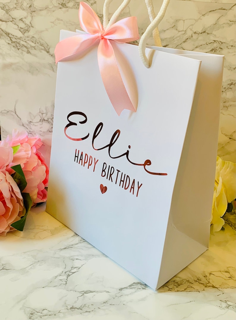May include: White gift bag with the name "Ellie" and "Happy Birthday" in rose gold script. A pink satin bow adorns the top, with white rope handles. A small red heart is below the birthday message. The bag is on a marble surface.