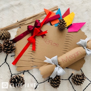 May include: A set of brown kraft paper crackers and flat cardstock shapes featuring the names Chloe and Megan in red script. The items are decorated with red, burgundy, and silver satin ribbons, colorful paper triangles, and pinecones.