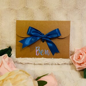 May include: A brown paper gift envelope tied with a vibrant blue satin ribbon. The name "Ben" is written in iridescent script on the front. Soft pink and cream-colored roses are arranged around the envelope.