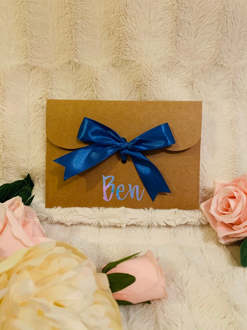 May include: A brown paper gift envelope tied with a vibrant blue satin ribbon. The name "Ben" is written in iridescent script on the front. Soft pink and cream-colored roses are arranged around the envelope.