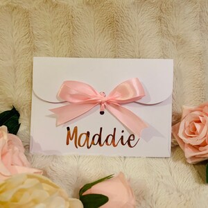 May include: A white gift card envelope with a pink satin bow. The name "Maddie" is written in a copper-colored script. The envelope is surrounded by pink roses and rests on a textured, cream-colored surface.