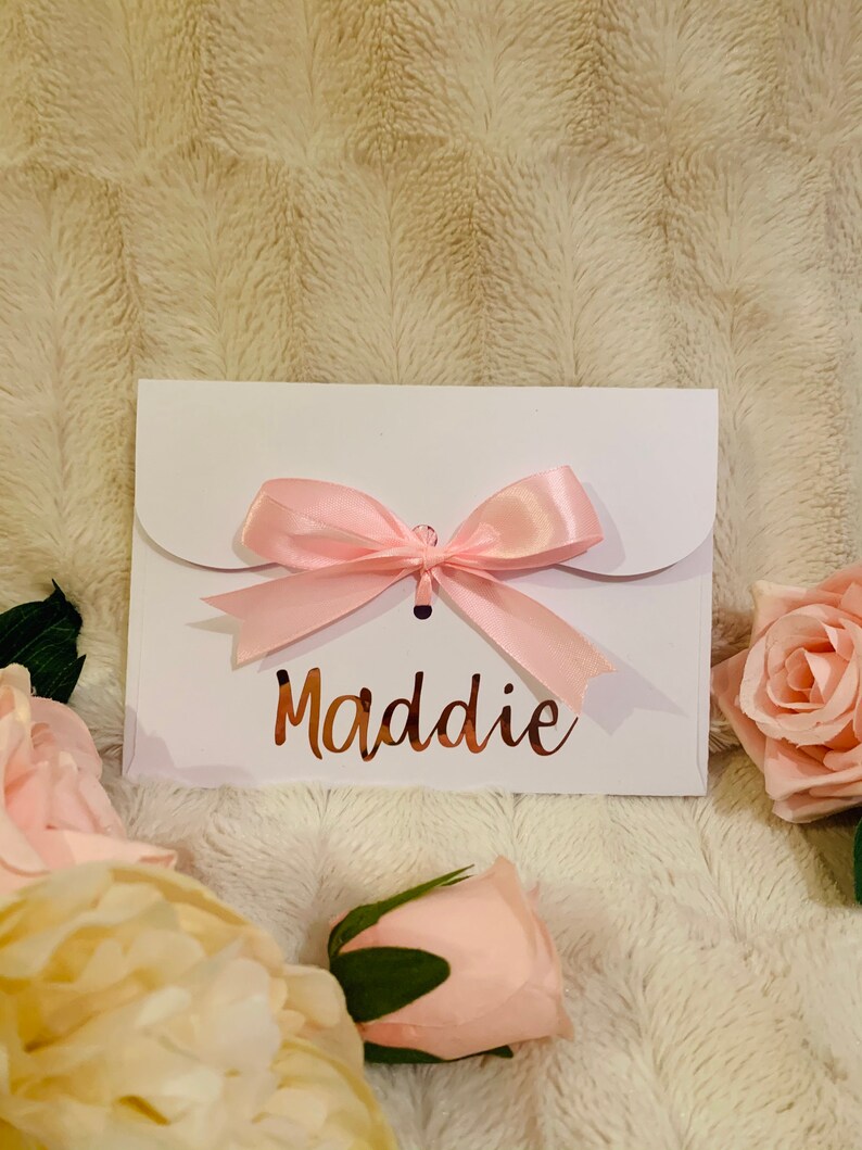 May include: A white gift card envelope with a pink satin bow. The name "Maddie" is written in a copper-colored script. The envelope is surrounded by pink roses and rests on a textured, cream-colored surface.