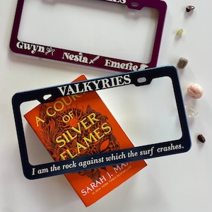 Puede incluir: Dos marcos de matrícula con la palabra "VALKYRIES" impresa en ellos. El marco superior es morado y tiene los nombres "Gwyn", "Nesta" y "Emerie" impresos en él. El marco inferior es azul y tiene un libro titulado "A Court of Silver Flames" de Sarah J. Maas, #1 New York Times Bestseller, sentado encima. El libro tiene una cita en la parte inferior que dice "Soy la roca contra la que las olas rompen."