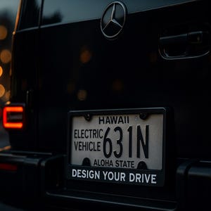 以下が含まれることがあります： 銀色のスリーポインテッドスターのエンブレムが付いた黒いメルセデス・ベンツ車の背面。ハワイのナンバープレートには「ELECTRIC VEHICLE 631N ALOHA STATE」と記載されており、「DESIGN YOUR DRIVE」と書かれた黒いホルダーに収められています。