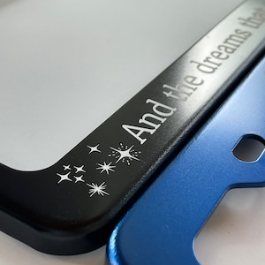 Peut inclure: Cadre de plaque d'immatriculation noir avec le texte "And the dreams they" et un motif d'&eacute;toiles blanches. Le cadre recouvre partiellement un cadre de plaque d'immatriculation bleu.