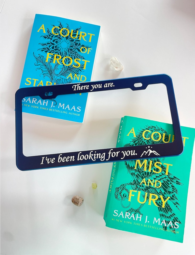 Peut inclure: Un cadre de plaque d'immatriculation bleu avec le texte "There you are." et "I've been looking for you." avec un graphique de montagne. Le cadre est &agrave; c&ocirc;t&eacute; de deux livres avec des couvertures bleu sarcelle pr&eacute;sentant un motif floral et le texte "A Court of Frost and Starlight" et "A Court of Mist and Fury" de Sarah J. Maas.