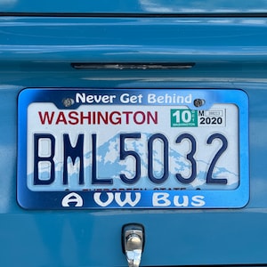以下が含まれることがあります： 上部に白い文字でNever Get Behind、下部にA VW Busと書かれた青い金属製のナンバープレートフレーム。このフレームは、BML5032という英数字と山のグラフィックが描かれたワシントン州のナンバープレートを囲んでいます。