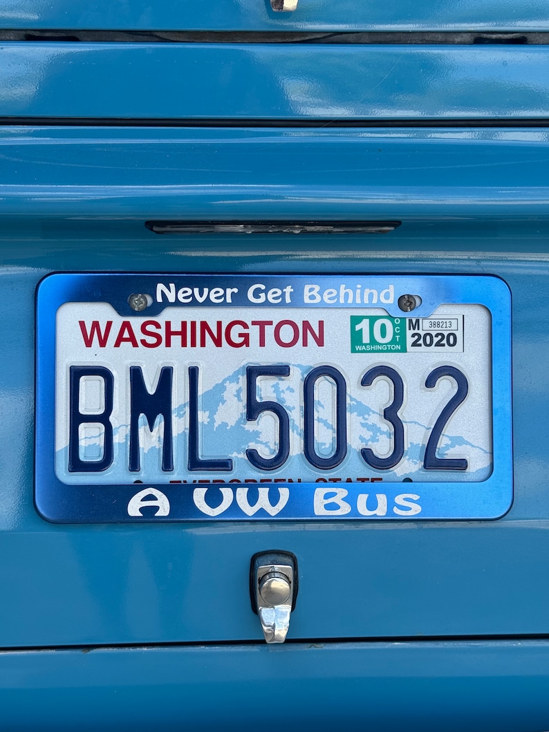 以下が含まれることがあります： 上部に白い文字でNever Get Behind、下部にA VW Busと書かれた青い金属製のナンバープレートフレーム。このフレームは、BML5032という英数字と山のグラフィックが描かれたワシントン州のナンバープレートを囲んでいます。