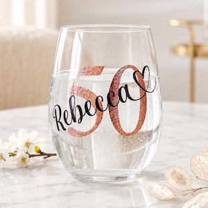 Op de afbeelding: Een helder, steelloos wijnglas met de naam Rebecca in zwarte cursieve letters, geplaatst over een groot, glinsterend ros&eacute;gouden getal 50. Het getal 50 is gestileerd met een hartvorm die in de nul is verwerkt voor een decoratief ontwerp.