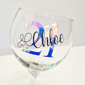May include: A clear glass goblet with a stem, featuring the number "21" in iridescent blue and pink, and the name "Chloe" in black cursive script. The glass is designed for beverages.