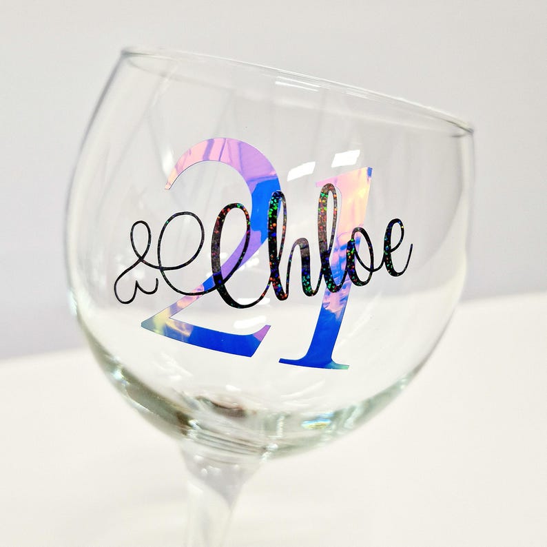 May include: A clear glass goblet with a stem, featuring the number "21" in iridescent blue and pink, and the name "Chloe" in black cursive script. The glass is designed for beverages.