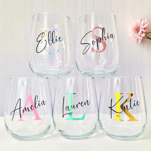 May include: Five clear glass wine glasses with personalized names and initial letters in colorful vinyl. The names are Ellie, Sophia, Amelia, Lauren, and Katie.