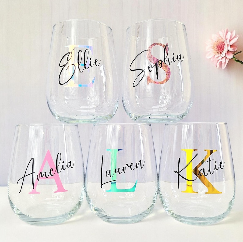 May include: Five clear glass wine glasses with personalized names and initial letters in colorful vinyl. The names are Ellie, Sophia, Amelia, Lauren, and Katie.