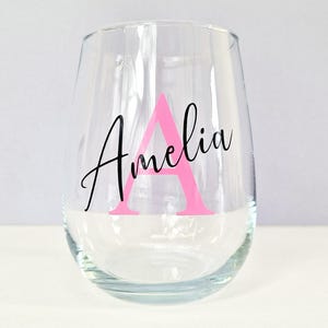 May include: A clear glass wine glass with the name "Amelia" written in black script with a pink letter "A" in the center.