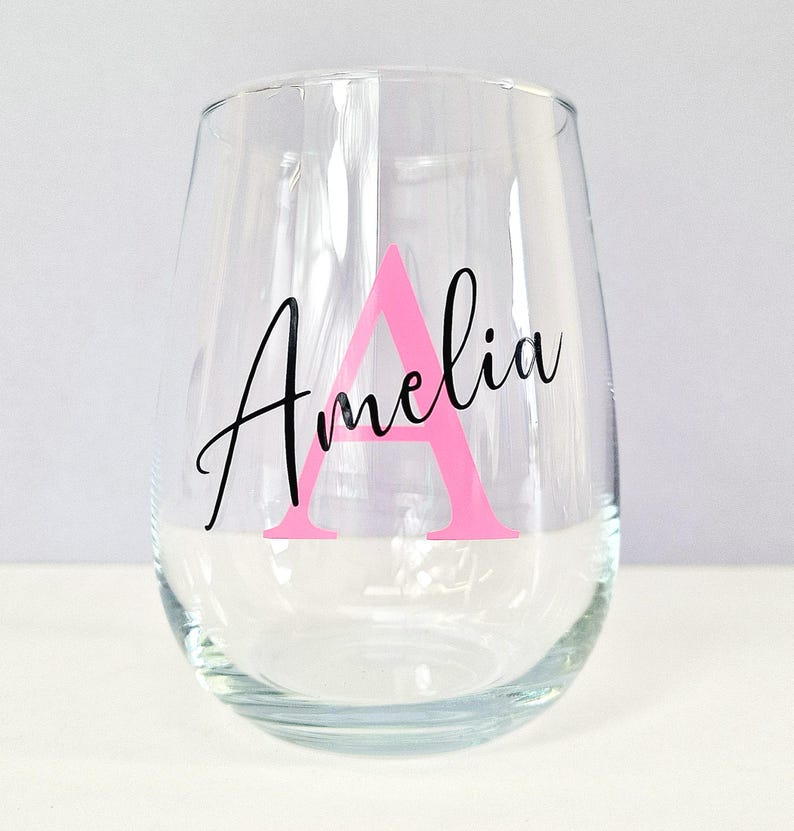 May include: A clear glass wine glass with the name "Amelia" written in black script with a pink letter "A" in the center.