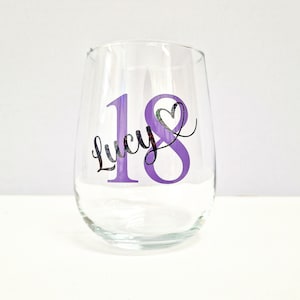 May include: A clear stemless wine glass with a purple "18" and the name "Lucy" in black script. A heart shape is incorporated into the "8". The glass is designed for a birthday celebration.