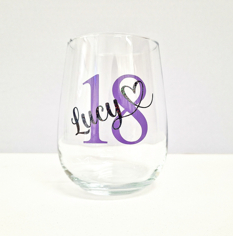 May include: A clear stemless wine glass with a purple "18" and the name "Lucy" in black script. A heart shape is incorporated into the "8". The glass is designed for a birthday celebration.
