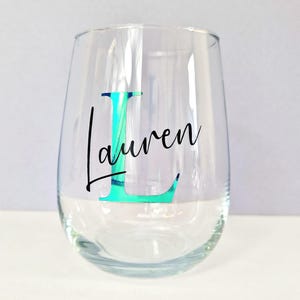 May include: A clear glass wine glass with a teal iridescent letter "L" and the name "Lauren" written in black cursive.