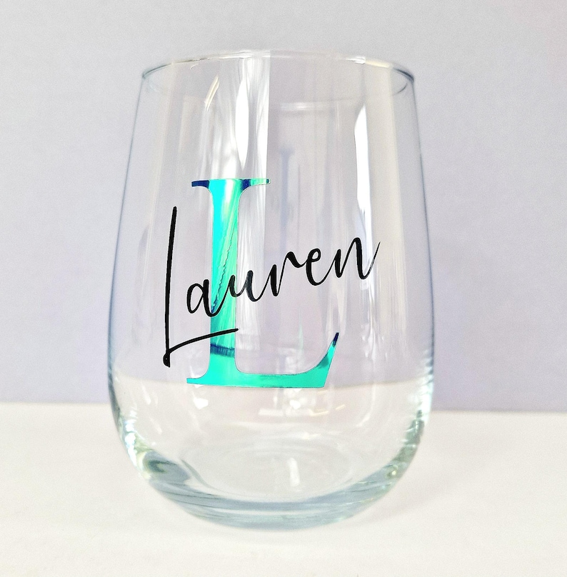 May include: A clear glass wine glass with a teal iridescent letter "L" and the name "Lauren" written in black cursive.