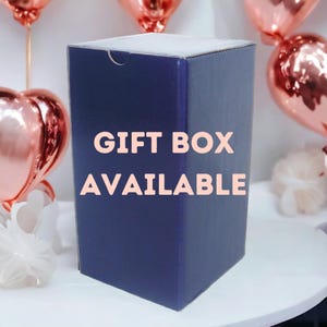 May include: A navy blue gift box with the text "GIFT BOX AVAILABLE" in a light pink font. The box is rectangular and has a white top. Rose gold heart-shaped balloons are in the background.