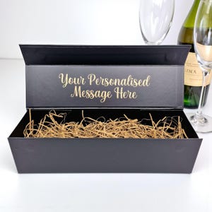 May include: A black gift box with an open lid, filled with brown packing straw. The box has gold text that reads "Your Personalised Message Here". A bottle of wine and two champagne flutes are in the background.