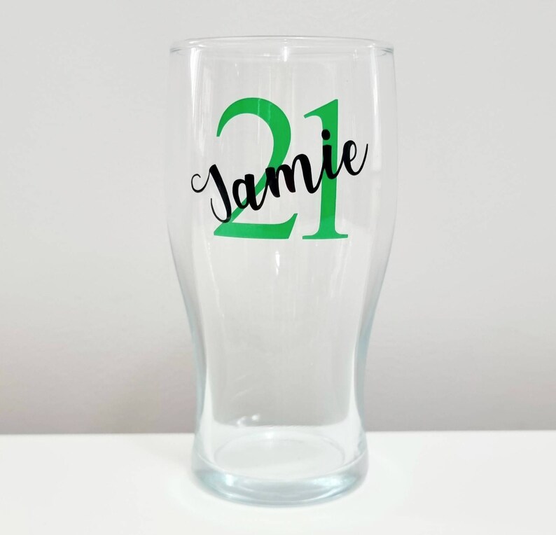 Puede incluir: Un vaso de pinta de vidrio transparente con una pegatina de vinilo verde y negro que dice "Jamie 21".