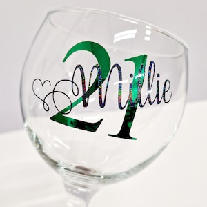 May include: A clear glass goblet with a green and black design. The number "21" is in green, with the name "Millie" in a glittery font. A heart and swirls are on the left side of the glass. The goblet is on a white background.