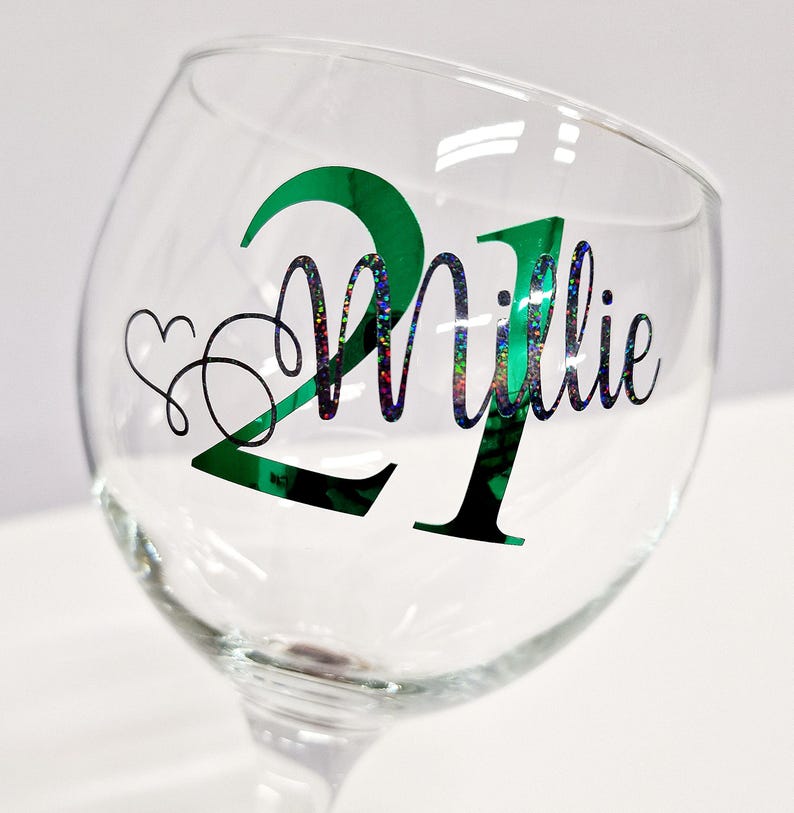 May include: A clear glass goblet with a green and black design. The number "21" is in green, with the name "Millie" in a glittery font. A heart and swirls are on the left side of the glass. The goblet is on a white background.