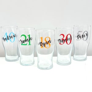May include: Five clear glass pint glasses with personalised names and numbers. The names are Andrew, Jamie, Ryan, Daniel, and Peter. The numbers are 40, 21, 18, 30, and 50. The numbers are in different colours.