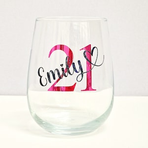 May include: A clear stemless wine glass with the number "21" in bright pink and the name "Emily" in black script. The glass is designed for a birthday celebration.