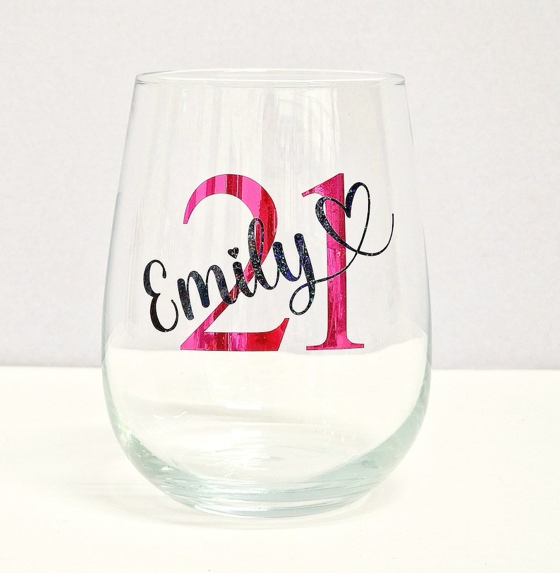 May include: A clear stemless wine glass with the number "21" in bright pink and the name "Emily" in black script. The glass is designed for a birthday celebration.