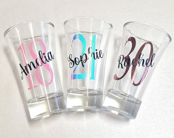 May include: Three clear glass shot glasses with personalized names and numbers. The shot glass on the left has the name Amelia and the number 18 in pink. The middle shot glass has the name Sophie and the number 21 in blue. The shot glass on the right has the name Rachel and the number 30 in brown.