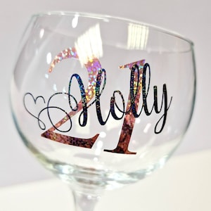 May include: A clear glass goblet with a stem, featuring the name "Holly" and the number "21" in a decorative font. The number "21" has a glittery, iridescent finish, and a heart-shaped design is next to the name.