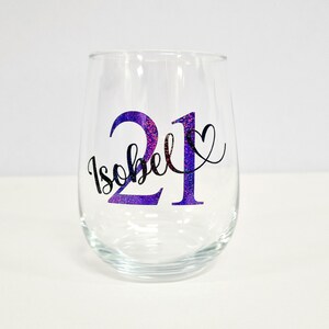 May include: A clear stemless wine glass with a purple glitter design. The design includes the number "21", the name "Isobel", and a heart. The glass is suitable for a birthday celebration.