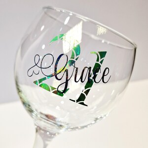 May include: A clear wine glass with the words "21 & Grace" in black script. The number "21" is decorated with a green and iridescent scale pattern. The glass is tilted, showing the stem and bowl.