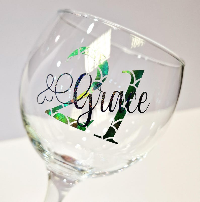 May include: A clear wine glass with the words "21 & Grace" in black script. The number "21" is decorated with a green and iridescent scale pattern. The glass is tilted, showing the stem and bowl.