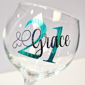 May include: A clear wine glass with the number "21" in teal and green glitter, and the name "Grace" in black script. The glass is tilted, showcasing the design on the front. The stemware is ideal for a birthday celebration.