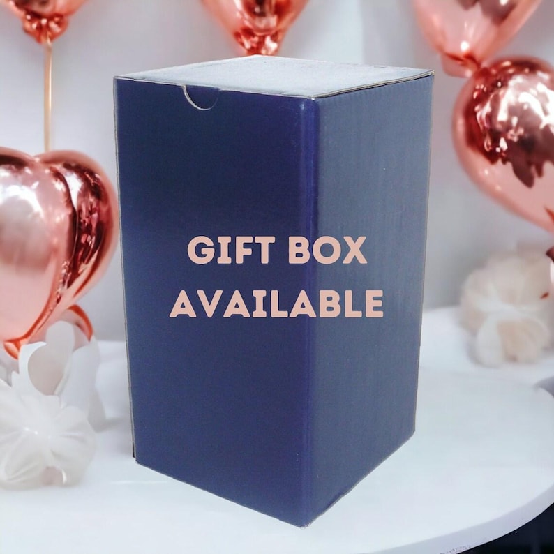Op de afbeelding: Een rechthoekige marineblauwe kartonnen geschenkdoos met een halfronde uitsparing aan de bovenrand. De tekst GIFT BOX AVAILABLE staat in lichtroze hoofdletters op de voorkant van de doos, die omringd is door ros&eacute;gouden ballonnen.