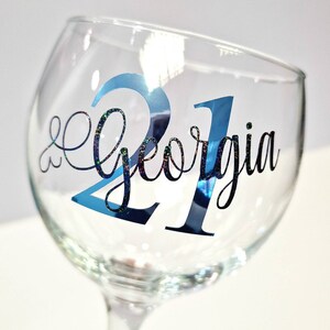 May include: A clear wine glass with the name "Georgia" and the number "21" in blue and black lettering. The number "21" has a shiny, reflective finish. The name "Georgia" is in a cursive font.
