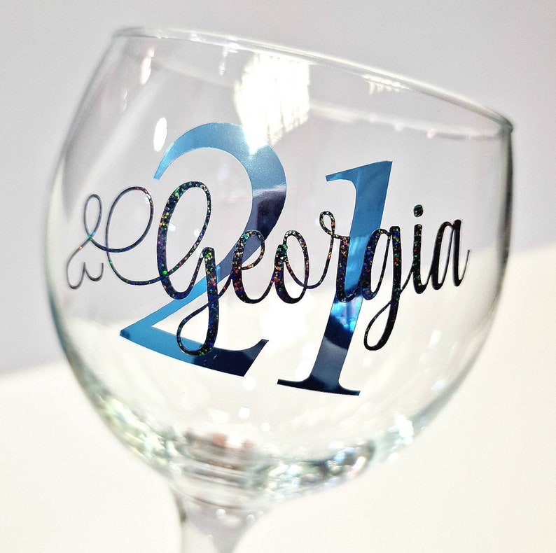 May include: A clear wine glass with the name "Georgia" and the number "21" in blue and black lettering. The number "21" has a shiny, reflective finish. The name "Georgia" is in a cursive font.