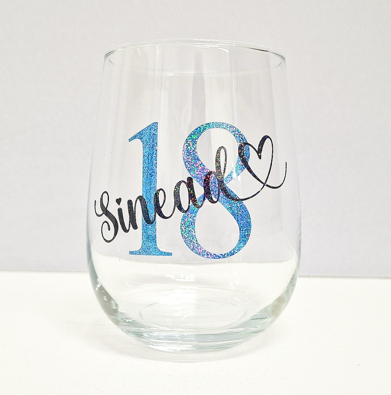 May include: A clear stemless wine glass with a blue glitter "18" and the name "Sinead" in black script. A black heart shape is next to the number 8. The glass is on a white surface.