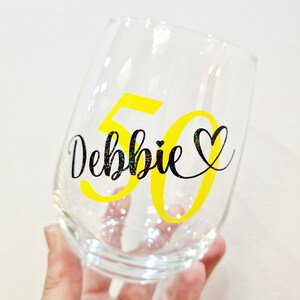 Op de afbeelding: Een helder, steelloos wijnglas met de naam Debbie in zwarte glitterende schrijfletters, vergezeld van een hartsymbool en het getal 50 in een groot, vet, felgeel lettertype dat achter de naam is geplaatst.