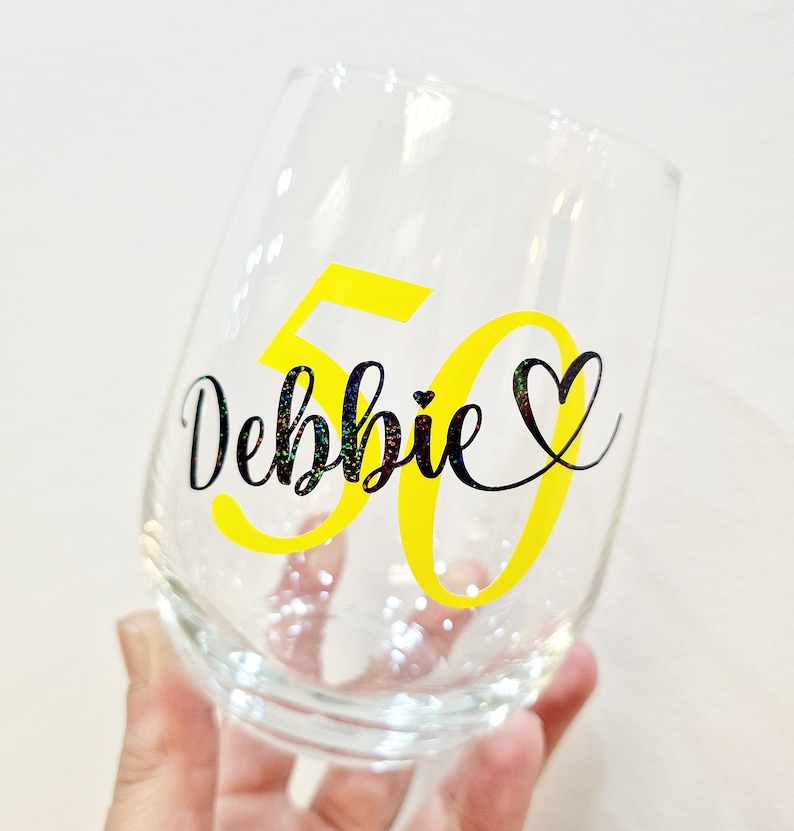 Op de afbeelding: Een helder, steelloos wijnglas met de naam Debbie in zwarte glitterende schrijfletters, vergezeld van een hartsymbool en het getal 50 in een groot, vet, felgeel lettertype dat achter de naam is geplaatst.