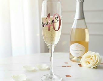 May include: A champagne flute filled with sparkling wine, featuring a rose gold "40" and the name "Laura" with a heart. A bottle of champagne and a white rose are in the background, along with scattered confetti.