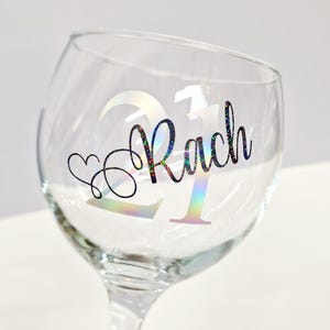 May include: A clear wine glass with a decorative design. The design includes the number "21" and the name "Rach" in a holographic, iridescent font. A heart shape is also included in the design.