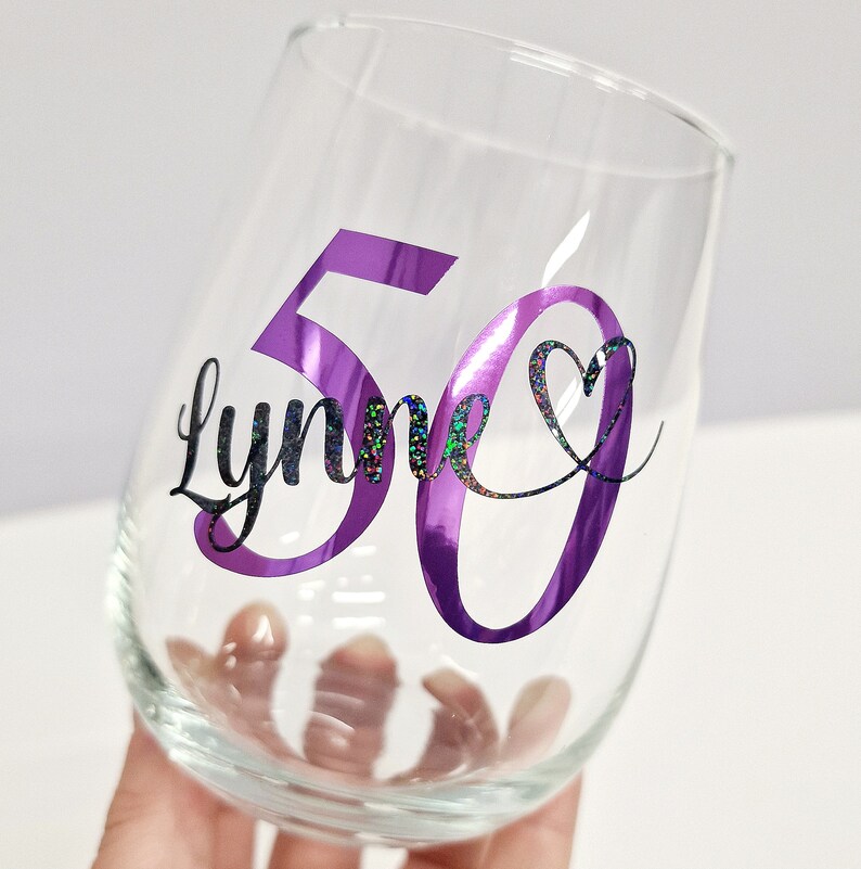 Op de afbeelding: Een helder, steelloos wijnglas met een groot paars getal 50 en de naam Lynne in zwarte, glinsterende schrijfletters. Een klein, zwart glinsterend hartsymbool bevindt zich rechts van de naam en maakt het ontwerp compleet.