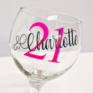 May include: A clear wine glass with a pink "21" and the name "Charlotte" in a decorative font. The text has a glittery, iridescent effect. The glass is on a white background.