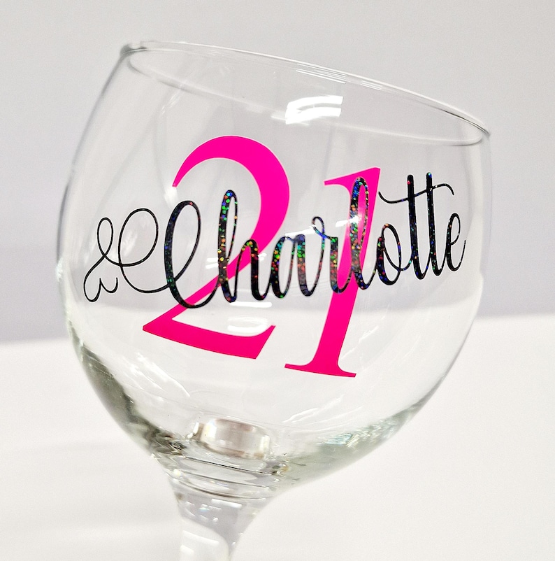 May include: A clear wine glass with a pink "21" and the name "Charlotte" in a decorative font. The text has a glittery, iridescent effect. The glass is on a white background.