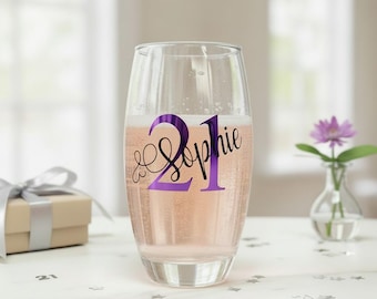 May include: A clear champagne flute filled with a pink beverage. The glass has a purple "21 & Sophie" design. A small gift box with a silver ribbon and a small vase with a purple flower are in the background. The setting is a celebratory theme.
