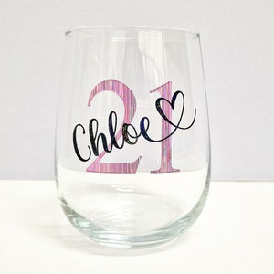 May include: A clear stemless wine glass with the number "21" in pink and a black heart. The name "Chloe" is written in cursive below the number. The glass is designed for a birthday celebration.